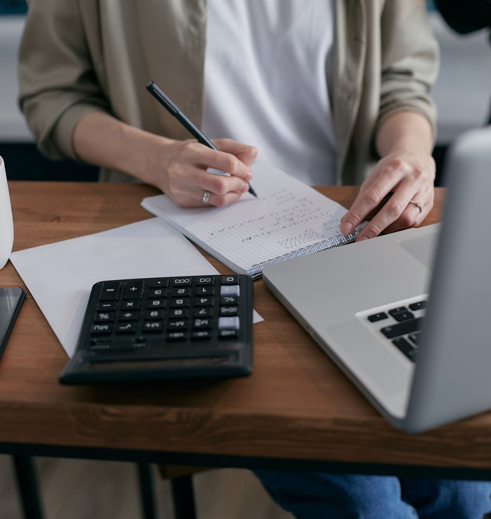 Mulher fazendo contas num caderno com calculadora e notebook ao lado