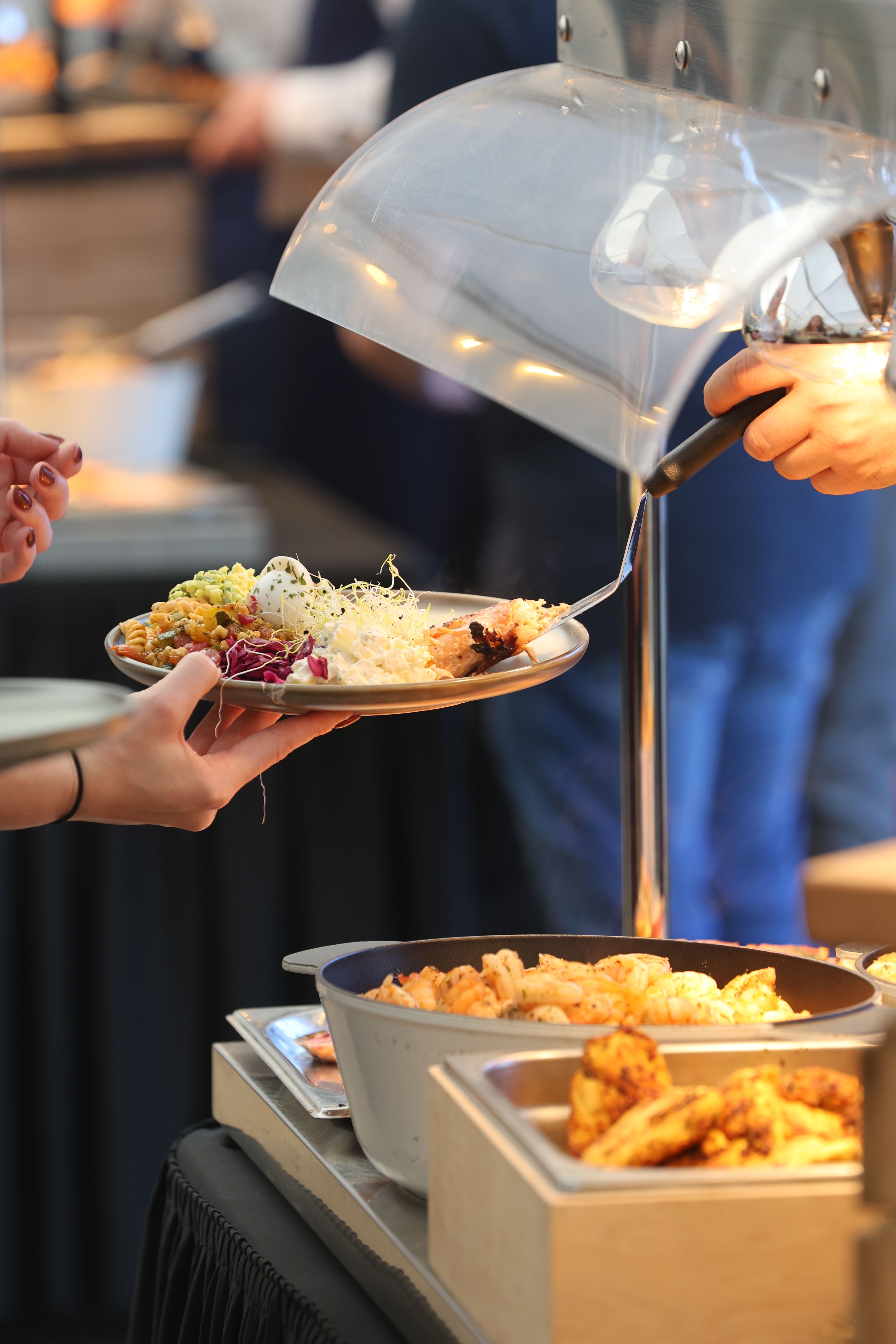 Funcionária de buffet servindo prato em evento, com estação de comidas quentes ao fundo